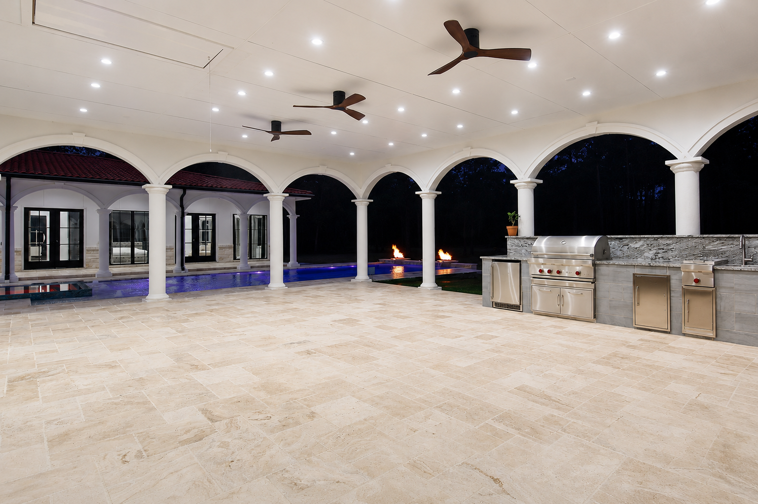 Covered veranda and outdoor kitchen at night with fire features on the pool